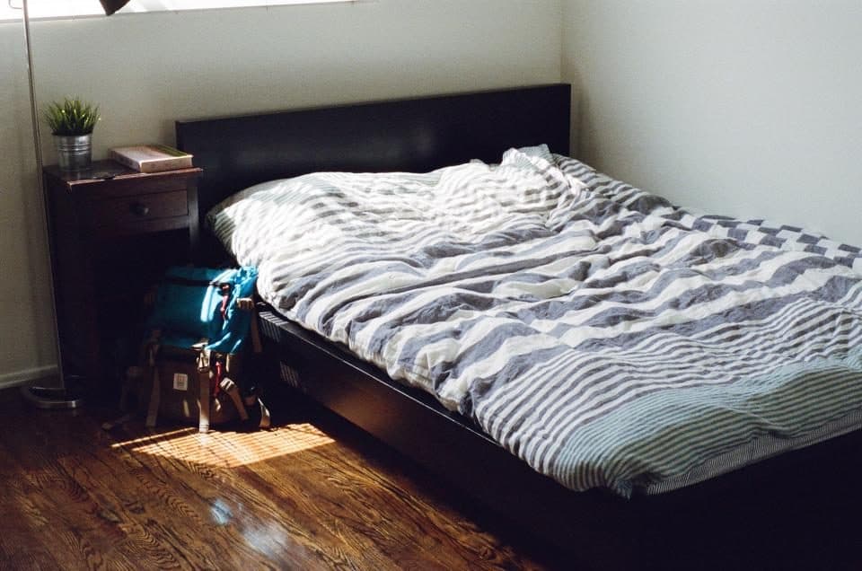 Un lit défait dans une chambre, un sac à dos posé au sol